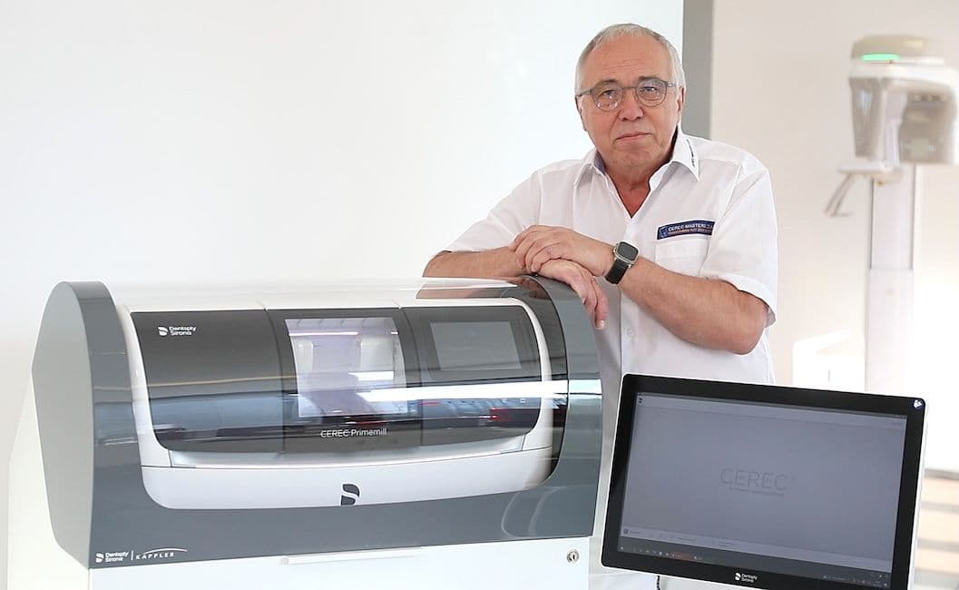 Ein Mann in einem Hemd mit Cerec-Masters-Club-Logo steht neben einer Cerec-Primemill-Fräseinheit und einem Monitor mit CEREC-Startbildschirm; im Hintergrund ist ein Scanner- bzw. Röntgengerät zu erkennen.