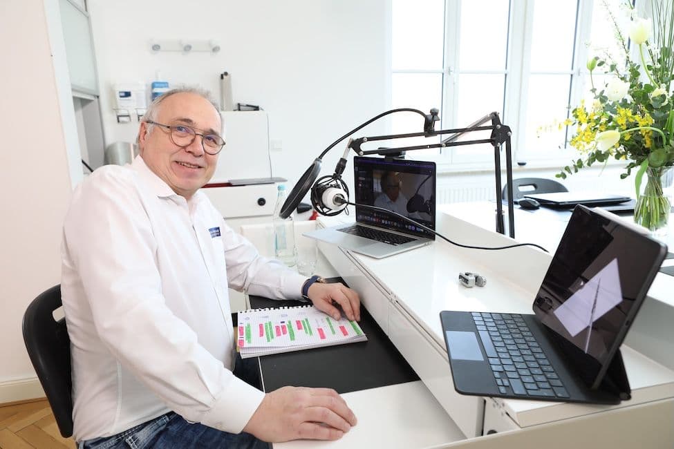 Dr. Andreas Kurbad blickt lächelnd in die Kamera, während im Hintergrund die Videokonferenz auf dem Laptop weiterläuft und ein Notizbuch mit farbig markierten Einträgen auf dem Tisch liegt.