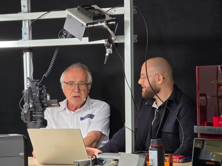 Zwei Männer bei einer Videoaufnahme in einem Studio mit Kamera, Mikrofon und Beleuchtungsausleger; im Hintergrund steht ein 3D-Drucker.