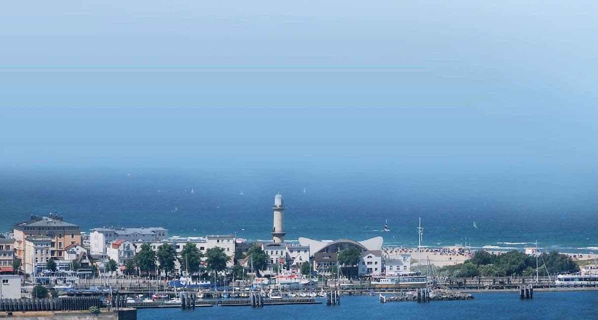 Blick über den Hafen von Warnemünde auf den Leuchtturm und das Gebäude mit dem geschwungenen Dach am Strand. Im Vordergrund liegen Boote an den Stegen, dahinter stehen Häuser und im Hintergrund ist die Ostsee mit Segelbooten zu sehen.