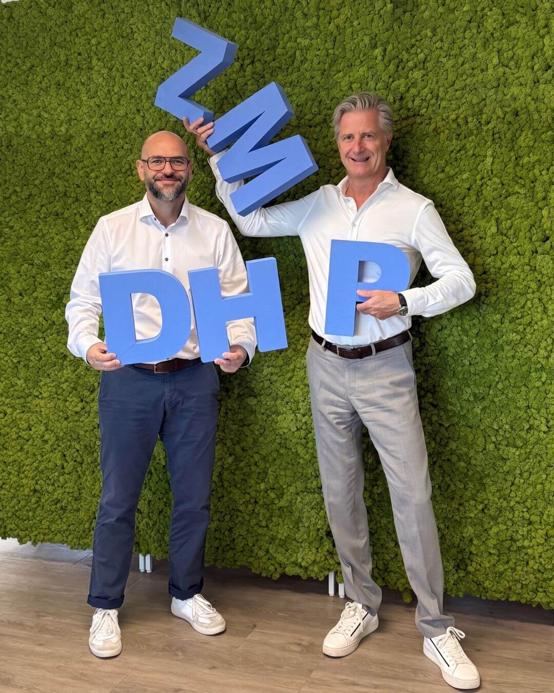 Zwei Männer in weißen Hemden stehen vor einer grünen Moosansicht und halten blaue 3D-Buchstaben in den Händen, die zusammen „DH ZMP" ergeben.